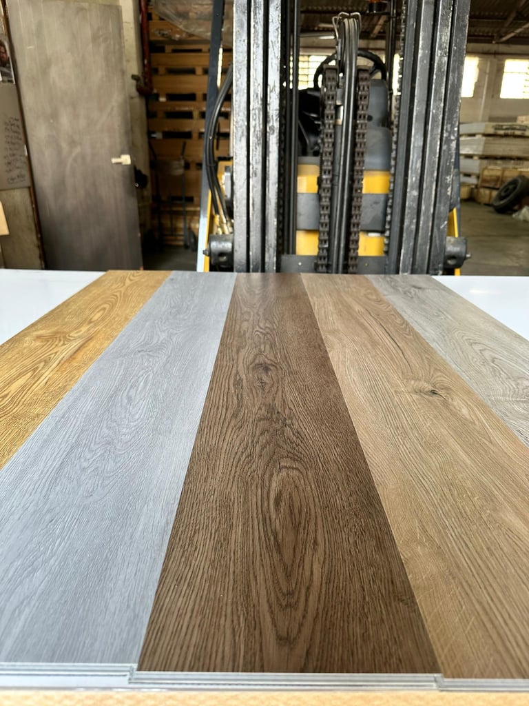 Three wood-look vinyl flooring samples displayed on a table showing different finishes from light to dark, with an industrial warehouse background