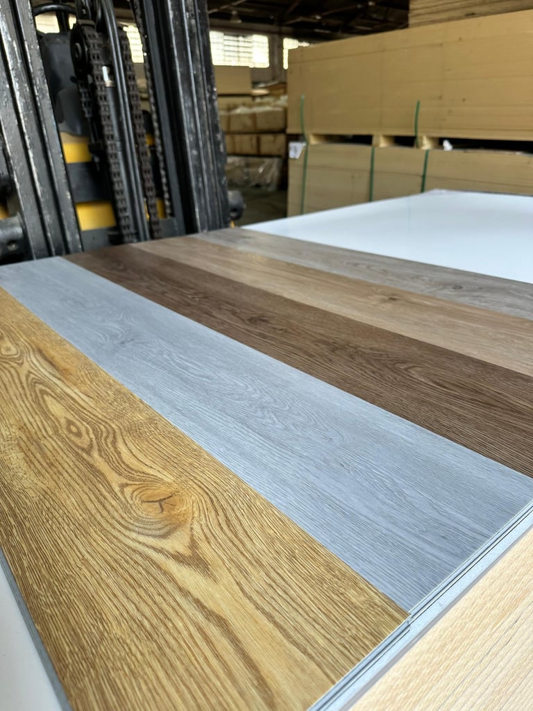 Sheets of wood-grain laminate flooring samples displayed in an industrial warehouse with stacked cardboard boxes and a forklift visible in the background